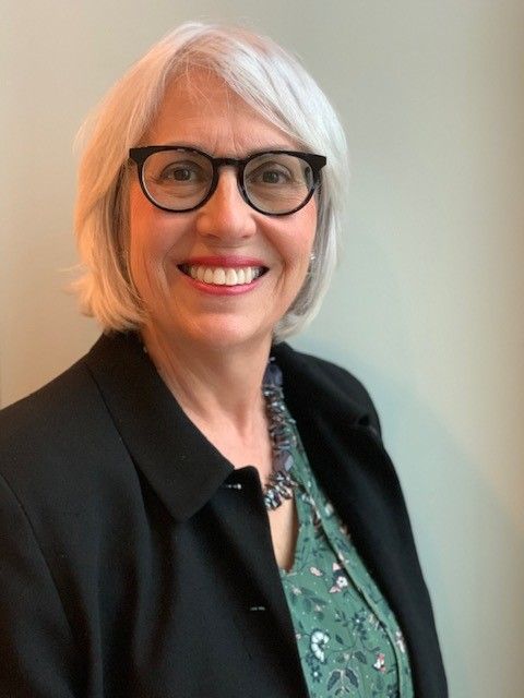 Smiling person with short white hair and glasses wearing a black blazer and patterned green top against a neutral wall.
