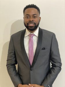 A person with a beard wearing a grey suit, white shirt, and light purple tie stands against a plain background.