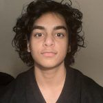 A person with dark, curly hair faces the camera with a neutral expression against a plain, light-colored background.