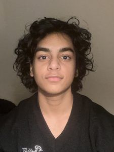 A person with dark, wavy hair and a neutral expression, wearing a black collared shirt against a plain light wall.