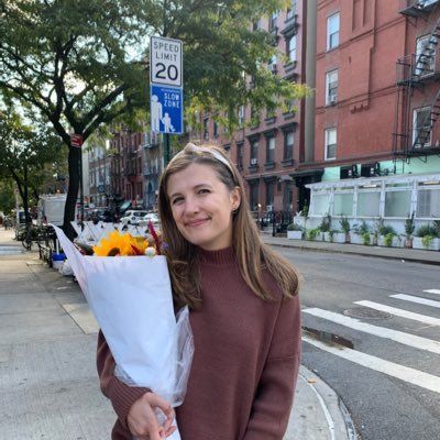 A person holding a bouquet of sunflowers on a city sidewalk next to a 20 mph speed limit sign and brick buildings.
