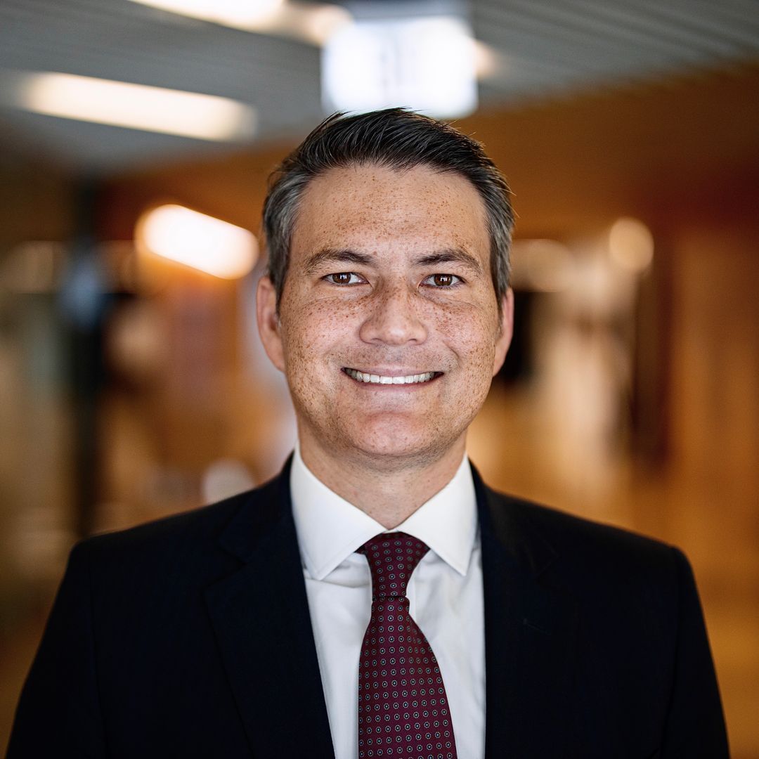 A smiling person in a dark suit, white shirt, and patterned red tie, standing in a softly blurred indoor office setting.