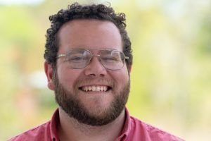 A smiling person with curly hair and glasses wears a red collared shirt, set against a soft, blurred outdoor background.