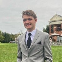 A smiling person in a light grey suit with a dark tie, standing outdoors on a green lawn near a large brick building.