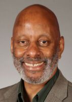 A smiling person with a beard, wearing a dark green shirt under a grey blazer, against a plain grey background.