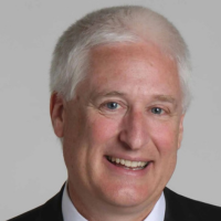 Headshot of a person with short white hair, wearing a dark suit and white collared shirt, smiling against a gray background.