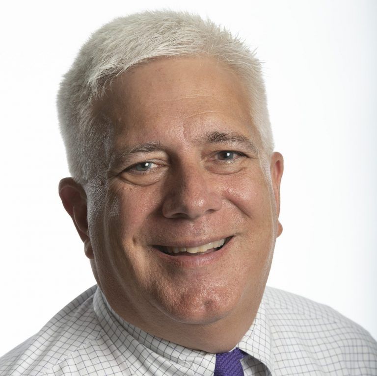 A smiling person with short, light hair wearing a white checkered shirt and purple tie against a plain white background.