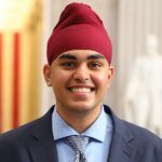 A smiling person wearing a maroon turban, light blue button-up shirt, patterned tie, and navy blue blazer.