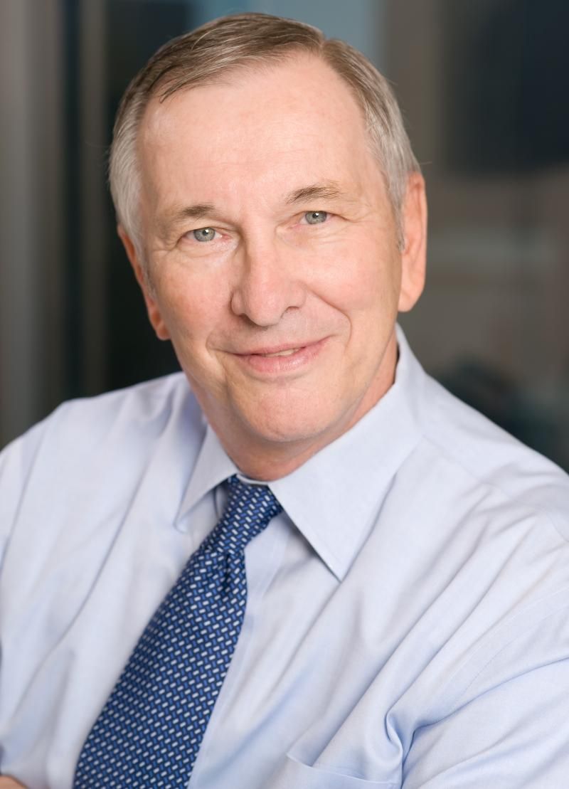 A professional headshot of a person wearing a light blue dress shirt and a blue patterned tie against a neutral background.