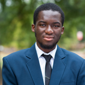 A person in a blue suit, white shirt, and black tie stands outdoors against a soft-focus green background.