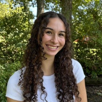 A smiling person with long curly brown hair, wearing a white shirt against a backdrop of green trees.