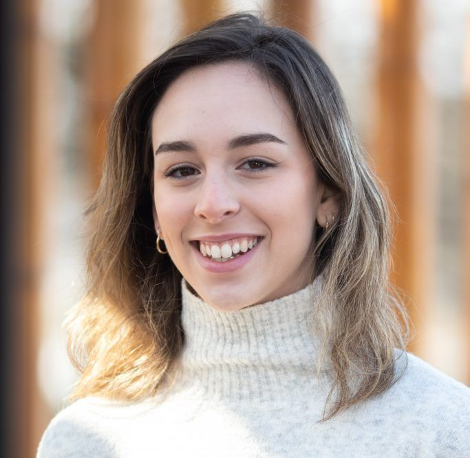 A smiling person with shoulder-length brown hair, wearing a cream-colored turtleneck sweater against a blurred background.