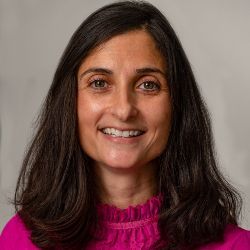 A smiling person with long, dark hair, wearing a vibrant magenta high-neck shirt against a neutral grey background.