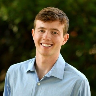 A smiling person wears a light blue button-down shirt against a soft, blurred green outdoor background.
