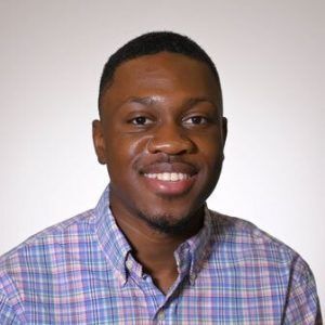 A person with a short haircut wearing a purple and blue plaid collared shirt smiles against a plain white background.
