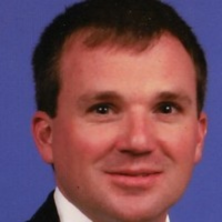 Headshot of a man with short brown hair, wearing a dark suit and white collared shirt, against a solid blue background.