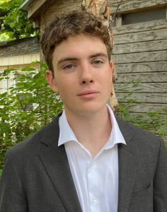 A person with curly hair wearing a white collared shirt and dark blazer stands outdoors in front of greenery and wood.