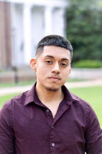 A person with short dark hair in a maroon collared shirt stands outdoors, looking toward the camera.