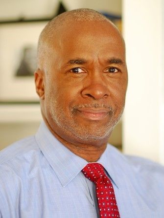 A person with a shaved head and light beard wearing a blue button-down shirt and red patterned tie, smiling softly.