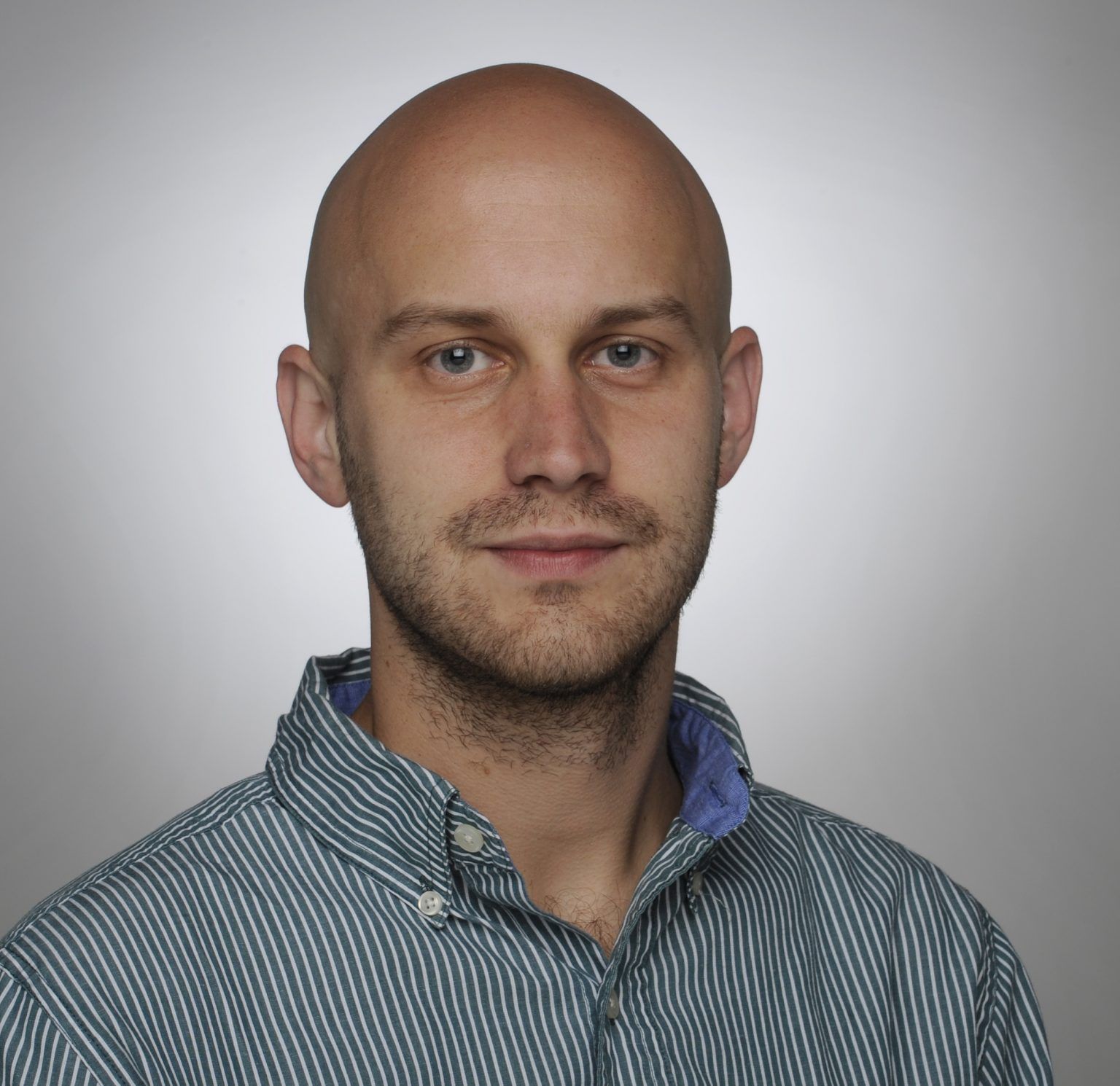 A headshot of a bald man with a short beard, wearing a green and white striped button-up shirt against a grey background.