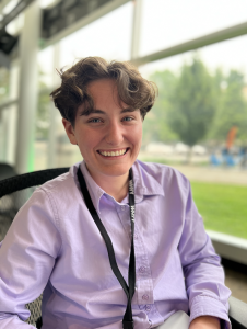 A person with wavy, dark hair wearing a light purple button-down shirt and a black lanyard, smiling at the camera.