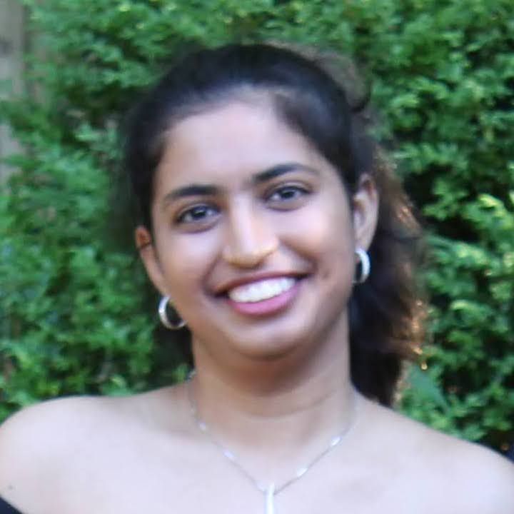 A person with dark hair and hoop earrings smiling broadly outdoors against a background of green foliage.
