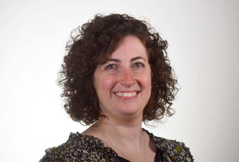A person with shoulder-length curly brown hair smiles warmly at the camera against a plain white background.