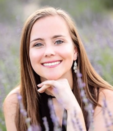 A smiling person with long brown hair, wearing dangling earrings, rests their chin on their hand in a lavender field.