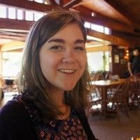 A smiling person with shoulder-length brown hair poses in a rustic, wood-paneled restaurant with large windows.