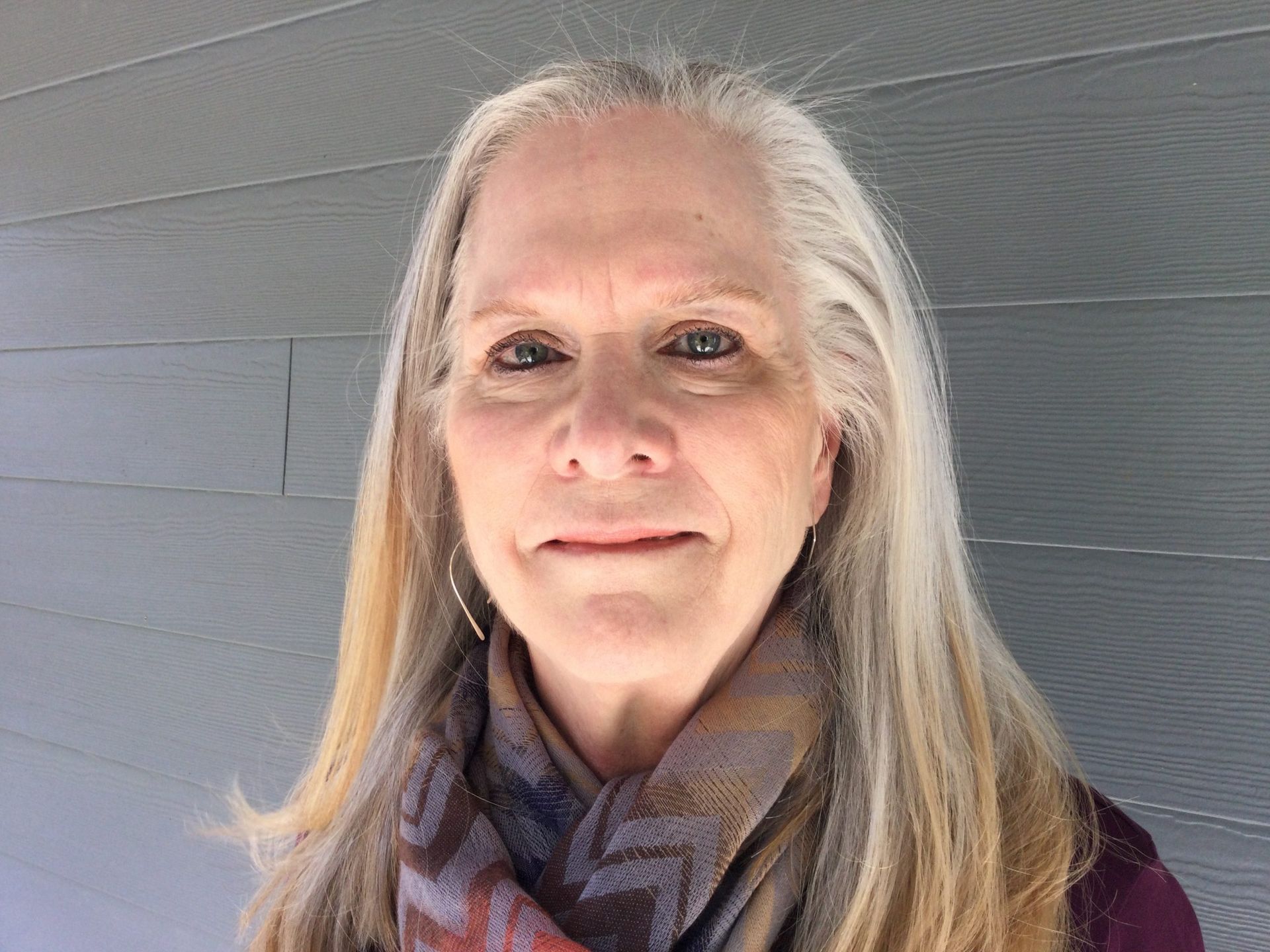 A person with long gray hair and a patterned scarf poses in front of a gray horizontal-paneled wall.