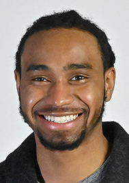 Headshot of a smiling person with dark hair and a beard, wearing a dark jacket against a white background.