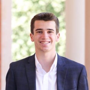 A smiling person wearing a dark blue patterned blazer and a white dress shirt, posed against a soft, out-of-focus background.