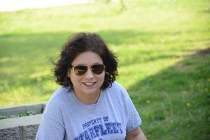 A person with dark, wavy hair and sunglasses smiles while wearing a grey 