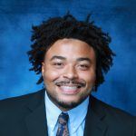 A person with curly hair smiling in a suit and light blue dress shirt against a mottled blue background.