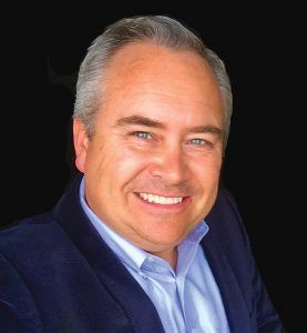 A smiling person wearing a blue button-down shirt and a navy blazer, centered against a solid black background.