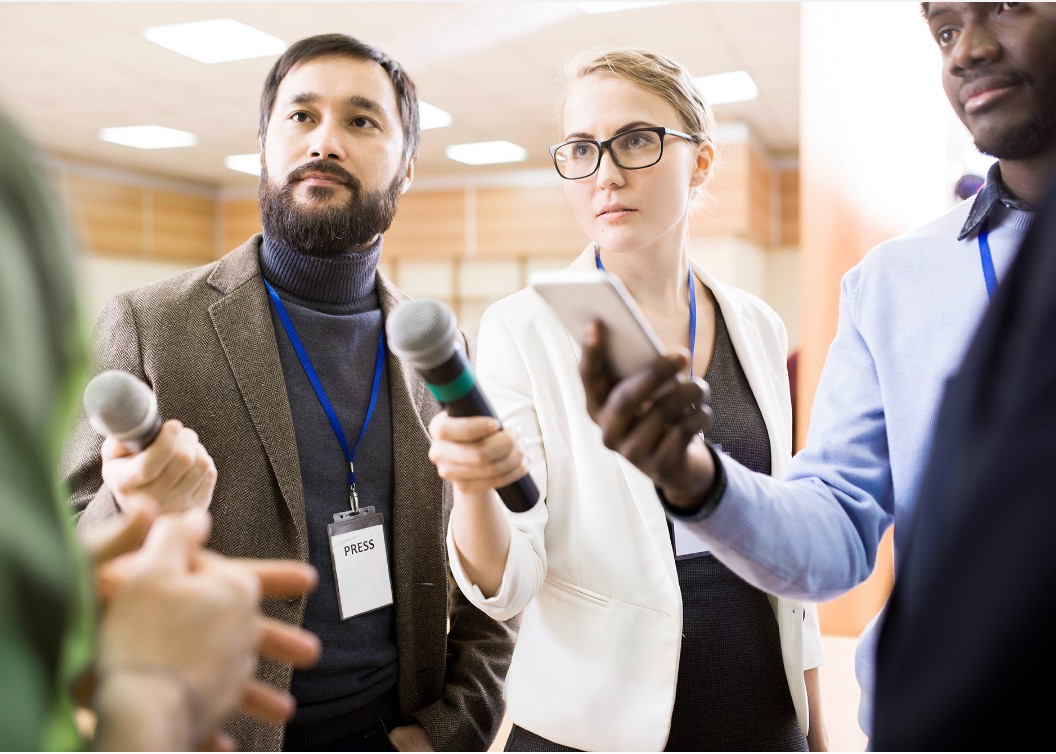 Three journalists hold microphones and a phone toward an unseen person, capturing an interview in a bright, indoor space.