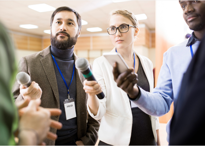 Three journalists hold microphones and a phone toward an unseen person, capturing an interview in a bright, indoor space.