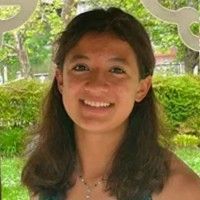 A smiling person with long dark wavy hair outdoors with green foliage and a white decorative fence in the background.