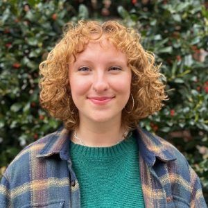A smiling person with shoulder-length curly reddish-blonde hair wearing a green sweater and a plaid button-up shirt.