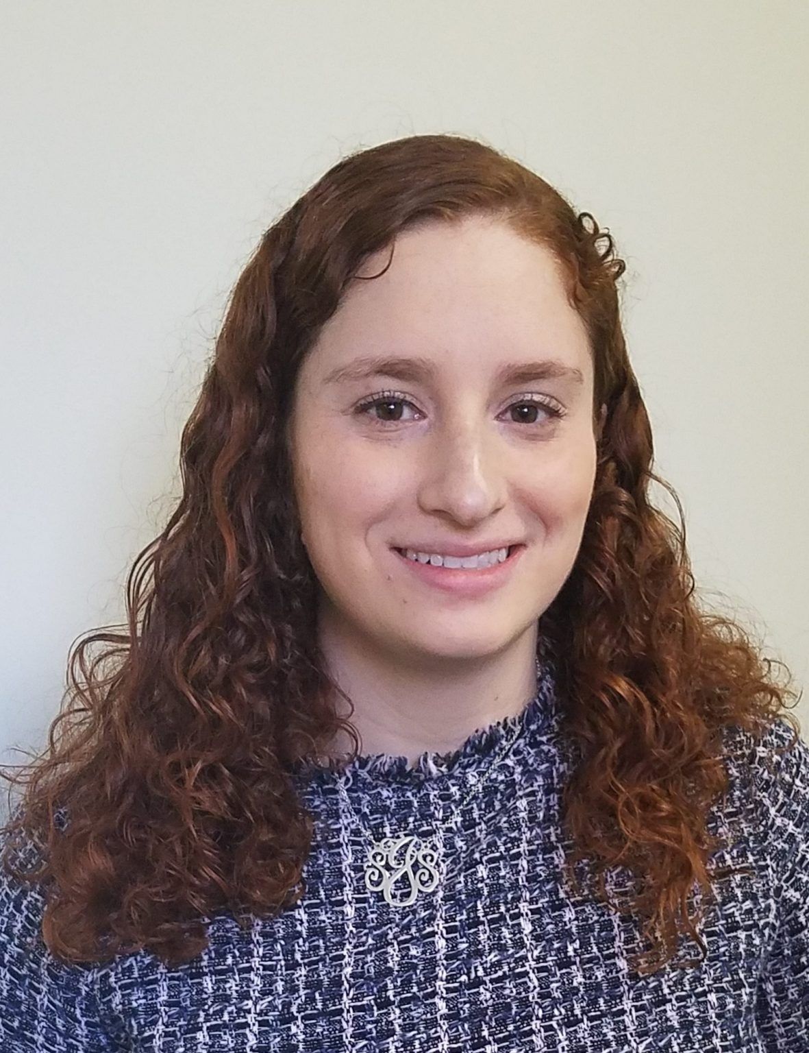 A person with shoulder-length curly reddish-brown hair, smiling, wearing a blue and white tweed shirt against a plain wall.