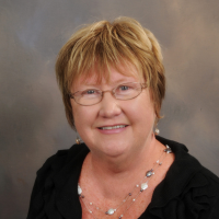A smiling person with blonde hair and glasses wearing a black top and a beaded necklace against a plain grey background.