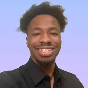 A person with dark hair and a goatee smiles at the camera, wearing a black collared shirt against a light blue background.