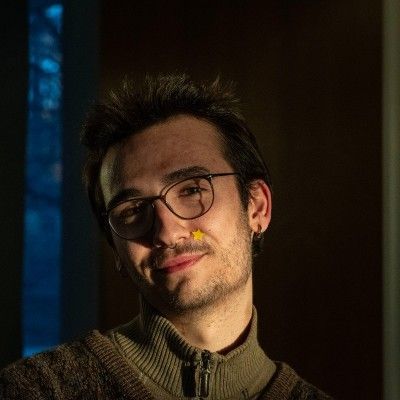 A person with glasses and a small yellow star sticker on their cheek, wearing a ribbed brown sweater in dim lighting.