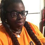 A person wearing glasses and an orange shirt smiles slightly while indoors, with their hair in two long braids.