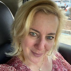 A blonde person with blue eyes, wearing gold hoop earrings and a cross necklace, smiles in a vehicle.