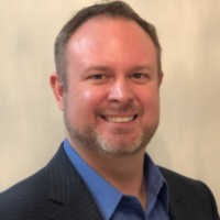 A man smiling, wearing a blue collared shirt under a dark navy blue blazer, against a plain, light-colored background.