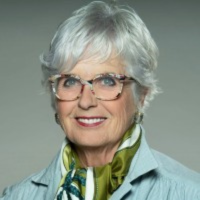 Smiling person with short white hair, wearing glasses, a green patterned scarf, and a blue collared shirt.