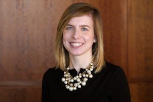 A smiling person with shoulder-length hair, wearing a black shirt and a statement pearl necklace against a wood background.