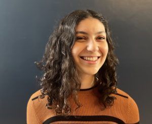 A smiling person with wavy dark hair wears a ribbed orange sweater with dark horizontal stripes against a dark background.
