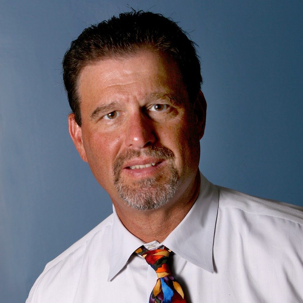 A person with short dark hair and a goatee, wearing a white shirt and a colorful patterned tie against a blue background.